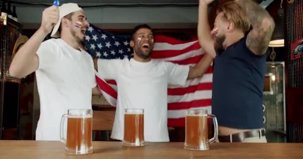 足球迷在体育酒吧庆祝球队的胜利 忠诚的足球迷支持他们的球队 拿着美国国旗 喝啤酒 看体育比赛的男人 — 图库视频影像