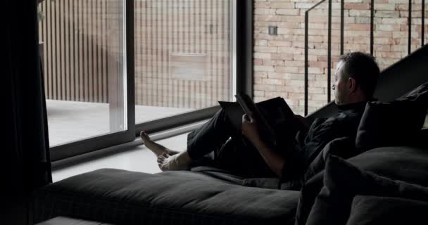 一个男人坐在黑色的沙发上看书 现代简约的客厅 有砖墙 大的滑动窗户和黑色家具 舒适的家具设计 奢华雅致的房间 — 图库视频影像