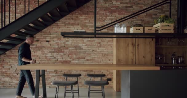 这个人坐在宽敞的现代木制厨房的椅子上 房间里有墙砖和黑色楼梯 房地产 豪华厨房 有木制岛屿和椅子 真正奢华的室内设计 — 图库视频影像