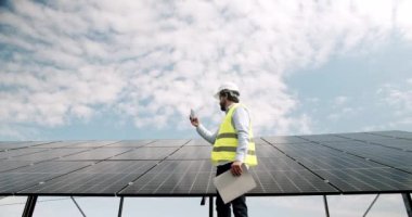 Erkek mühendis, güneş çiftliğinde akıllı telefondan konuşuyor. Düşük açılı üniformalı adam güneş enerjisi istasyonunda güneşli bir günde fotovoltaik panellerin yanında yürüyor ve bulutlu mavi gökyüzüne yapılan çağrıya cevap veriyor..