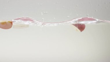 Temiz suya düşen elma parçaları. Beyaz arkaplanlı şeffaf temiz suyun yavaş hareketli durağan görüntüsü ve olgun kırmızı elma parçalarından dökülen parçacıklar.