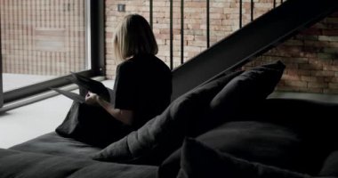 Bir kadın siyah kanepede oturuyor ve kitap okuyor. Modern Minimalist oturma odası, tuğlalar, büyük sürgülü pencereler ve siyah mobilyalar. Moda mobilyalar..