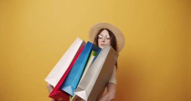 Female shopaholic with colorful paper bags. Static shot of happy young woman in glasses checking and showing shopping bags then touching hat while standing against yellow background