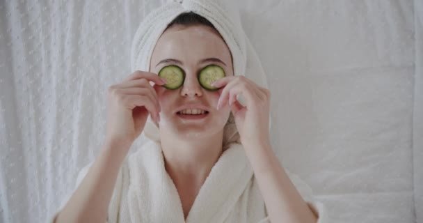 穿着浴衣 头上蒙着毛巾 头上罩着黄瓜 为新鲜清洁健康的护肤理念做广告的有趣而快乐的年轻女性 拿着有机温泉面具的女人笑着看着摄像机 — 图库视频影像
