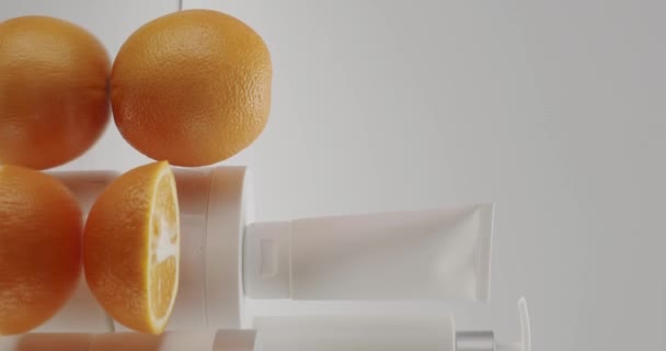 スタジオの白い背景に化粧品のための新鮮なオレンジ色の果物を持つ白いボトルとチューブ 天然成分 スクラブ 強壮剤 ボディケアに基づく化粧品油 ストーリーの垂直ビデオ — ストック動画