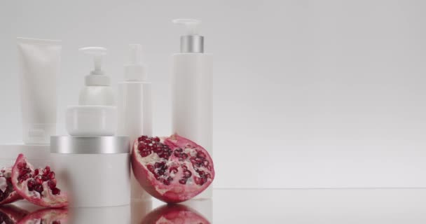 スタジオの白い背景に化粧品のためのザクロの果物と白いボトルとチューブ 天然成分に基づく化粧品油 スクラブ 強壮剤 ヘアケアのための天然化粧品生産 — ストック動画