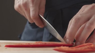 Front view of the young chef cut thin slices of a piece of red pepper with a knife on the cutting board. Front view of a young chef cutting thin slices of a piece of red pepper. Vegetables up close.