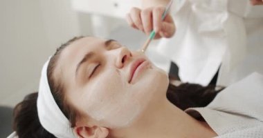 Güzellik uzmanı kozmetik ürün jel soyma maskesini spatulayla, cilt bakımıyla uyguluyor. Yüz yenileme konsepti. Genç kadın kozmetik ilaç tedavisi görüyor, yakın plan..