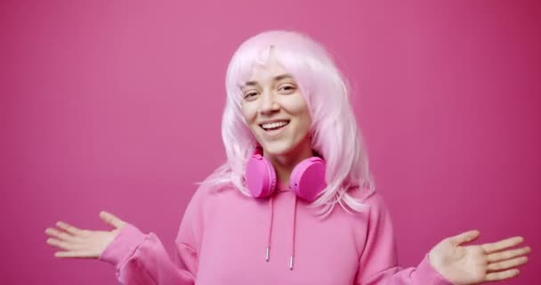在粉红的背景下庆祝成功的同时 身着帽衫 头戴无线耳机 头戴明亮的拳头 面带微笑地看着相机的年轻女性的静态照片 — 图库视频影像