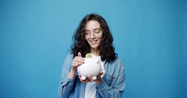 一个年轻的女人手里拿着一个存钱箱 手里拿着一个孤立的蓝色背景的钱 可爱的钱箱 Childs Coin Bank 可爱的Piggy银行钱箱 — 图库视频影像