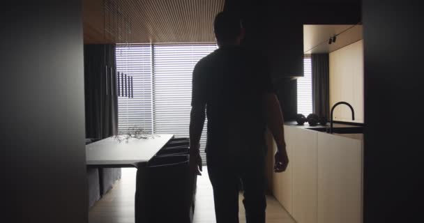 一个男人从窗户往外看 看到一间有现代化厨房内部的公寓 这个男人穿过厨房 停在窗前 带有木料和简约设计的现代餐厅 — 图库视频影像