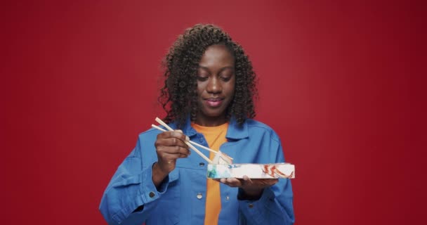 カメラと笑顔を見ているアフリカ系アメリカ人女性は 隔離された赤い背景で箸で寿司を食べる準備をしている 日本料理 寿司ロール アジアのストリートフード — ストック動画