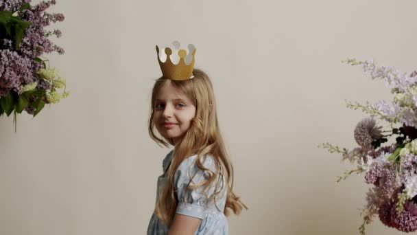 7岁的小女孩穿着浅蓝色的衣服 头戴金色的王冠 站在工作室的白色背景上看着摄像机 小金发美女脸 — 图库视频影像