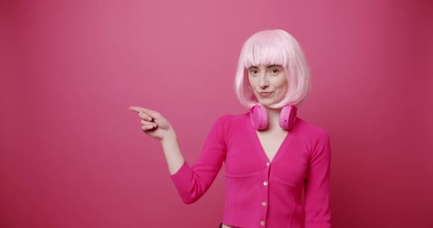 时髦的年轻女性指指点点 身着时髦服装 头戴无线耳机 头戴明亮头发 指着粉红背景微笑的友善年轻女子的静态照片 — 图库视频影像