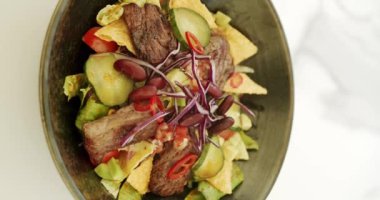 Etli salata, fasulye, vişneli domates, salatalık, kırmızı biber, kırmızı soğan, lahana salatası, beyaz arka planda dönen büyük tabakta nachos cipsi. Taco salatası. Kapatın. Dikey video.