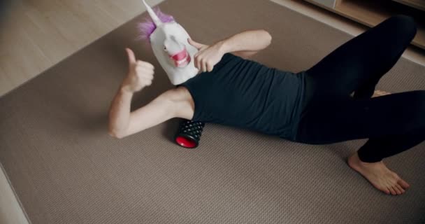 一个戴着独角兽面具的有趣男人在家里用滚子做运动来放松自己 在家里的滑稽健身 午餐休息时背痛减轻了 在家进行自我按摩的工具 — 图库视频影像