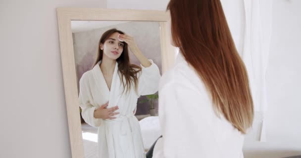 护肤身体护理 在家进行护理和清洁 漂亮的女人穿着白色浴衣 在家里的大镜子里 拿着棉垫 去除杂质 洗净她完美的皮肤 — 图库视频影像