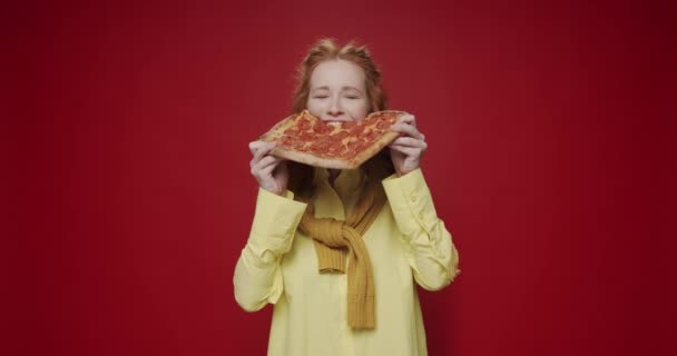 美丽饥饿的女孩吃着辣香肠披萨 在红色孤立的背景上享受和微笑 饥饿的女人喜欢吃比萨 吃一片好吃的意大利辣披萨 — 图库视频影像