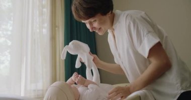 Neşeli gülümseyen anne yeni doğmuş bebekle oynuyor, pelüş tavşan oyuncakları gösteriyor ve bebek odasında bebekle konuşuyor. Mutlu ebeveynlik kavramı