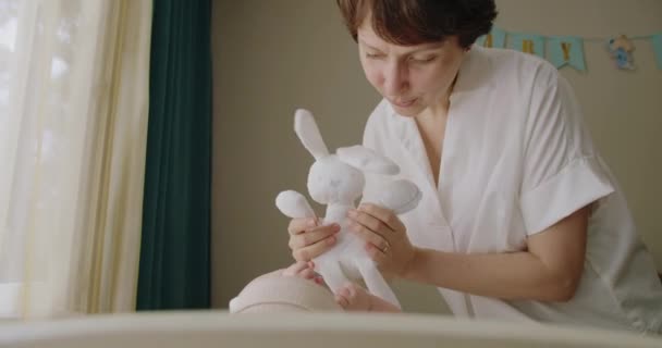母親は柔らかいおもちゃのウサギを手に持って 子供の部屋で赤ちゃんと遊んでいます 素敵な新生児を楽しんでいる遊び心のある女性 ハッピー — ストック動画