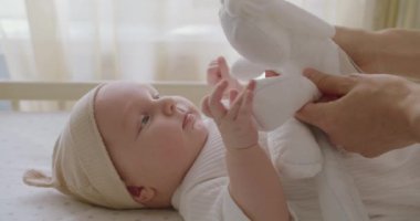 Beyaz tulum içinde beşikte yatan yeni doğmuş şirin bir bebek. Anne kadın, yumuşak oyuncağı elinde tutuyor ve bebek odasındaki güzel bebekle oynuyor..