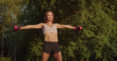 Atletik genç bayan, dışarıda halterlerle egzersiz yapıyor. Spor kıyafetli kadın yaz parkında sabah sporu yapıyor..
