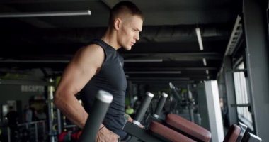 Sportif genç adam modern spor salonundaki eğitim aletleri üzerinde egzersiz yapıyor ve kameraya bakıyor. Sporcunun fitness merkezinde antrenmanı var. Spor ve sağlıklı yaşam tarzı kavramı