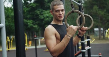 Spor sahasında durup kameraya bakan genç bir sporcunun portresi. Yakışıklı sporcu jimnastik yüzükleriyle egzersiz yapıyor..