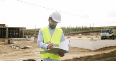Male engineer in uniform and white helmet makes notes in notebook at work. Verification of works on the territory of the construction site. Industrial Building Construction Site.