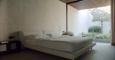 Yatak odasının içinde rahat çift kişilik yatak, yastıklar ve yatak odaları cam duvarlı, açık perdeli ve bahçe manzaralı modern yazlık evde.