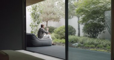 Genç adam modern kır evinin terasında rahat bir fasulye koltuğu üzerinde oturuyor, kahve içiyor ve doğanın tadını çıkarıyor. Düşünceli erkek dışarıda bir fincan çayla dinleniyor.