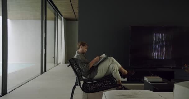 年轻人坐在舒适的椅子上 在有现代简约室内装饰和玻璃墙的客厅看书 — 图库视频影像