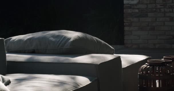 コテージハウス近くのテラスのラウンジゾーン 枕付きの快適なソファー 屋外の灰色のソファーの木の影 レストコンセプト — ストック動画