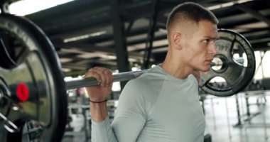 Ciddi bir sporcunun portresi ağır halterleri omuzlarında tutuyor ve spor salonunda kameraya bakıyor. Sporcu adam spor aletleriyle ağırlık çalışması yapıyor.