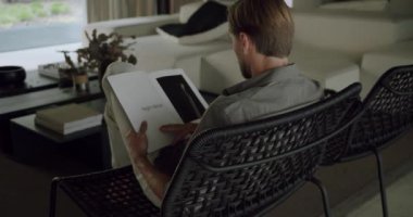 Rahat koltukta oturan, dergi okuyan ve modern apartmandaki dinlenme odasında dinlenen genç adamın arkası. Boş zamanlarında evde kitap rahatlığı olan bir erkek.