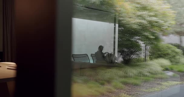 快適な椅子に座ってガラスの壁の背後にある豪華なアパートで本を読む男 ガラス ドアか窓が付いている現代コテージの家の中の雑誌と休む男性 — ストック動画