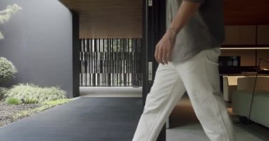 Manzaralı adam, modern yazlık evin terasındaki oturma odasından çık ve yaz sabahının tadını çıkar. Erkek, açık cam kapılı, yazlık evde duruyor.