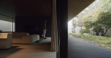 Şık giyinmiş genç adam terasta oturma odasından bahçeye bakıyor ve dışarıda sabahları eğleniyor. Modern tatil evinin verandasında cam panoramik sürgülü kapısı olan bir erkek.