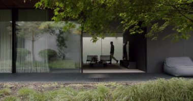Modern yazlık evin oturma odasında bir adam yürüyor. Açık cam sürgülü kapısı olan bir yazlık evin içi ve dışı modern mobilyalarla dolu.