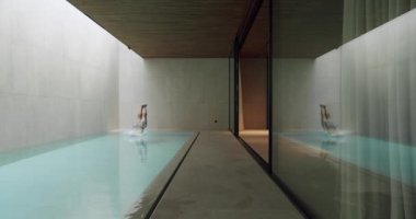 Atletik adam havuza atlıyor, suya dalıyor ve yüzüyor. Lüks spa otelinde dinlenme ve dinlenme. Camdan kayan panoramik kapısı ve modern iç mekanı olan bir daire..