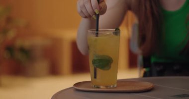 Masadaki kafede oturan, saman tutan ve sarı kokteyli buz ve nane yaprağıyla karıştıran bir kadın. Kadın soğuk limonata içiyor, tatlı bir tadı var.