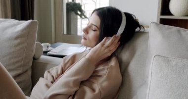 Mindful young happy woman wearing wireless headphones, listening relaxing music, resting on comfortable sofa, enjoying interesting audio book or affirmations, spending leisure indoors