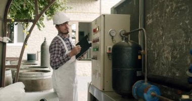 Erkek şarap imalatçısı, içeceklerin kalitesini içeride kontrol ediyor ve günlüğe tarihleri yazıyor. Alkol üretim kavramı. Şarap fabrikasındaki üretim ekipmanlarının kalitesini kontrol eder.