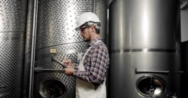 Erkek şarap imalatçısı, içeceklerin kalitesini içeride kontrol ediyor ve günlüğe tarihleri yazıyor. Modern şarap imalathanesi veya şarap imalathanesi. Şarap fermantasyonu için büyük çelik fıçılar..