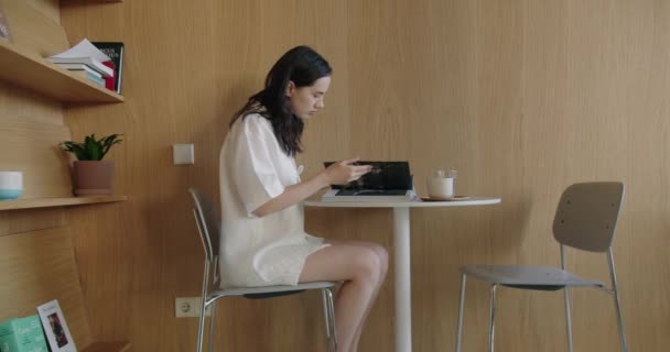カフェテーブルに座っている若い女性は ラテと朝食の間に本を読んでいます モダンなインテリアのコーヒーショップで休んでいる間 女性学生のフリッピング雑誌のページ — ストック動画