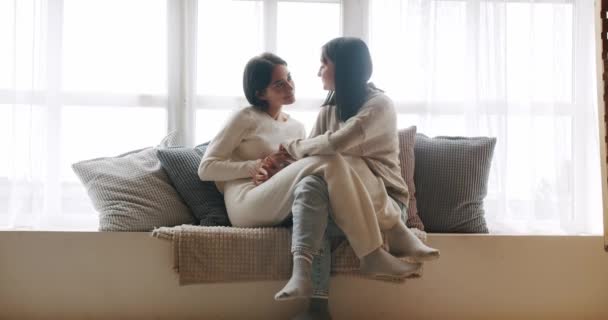 Lgbt和同性恋妇女在家里 同性恋夫妇享受的肖像 一对年轻的女同性恋夫妇在家里明亮的客厅靠窗坐在沙发上 一边微笑 一边轻轻地触摸着双手 — 图库视频影像