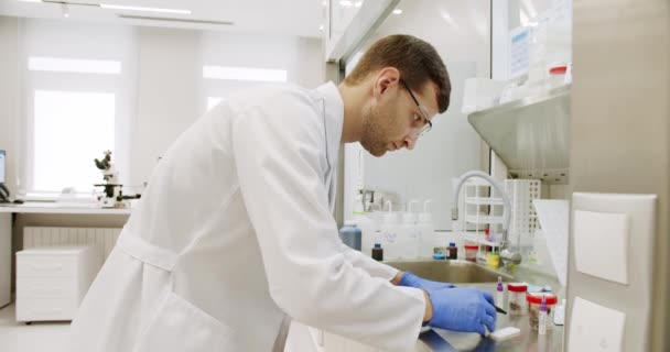 現代医学研究所 手袋とメガネで白いローブの男性の手持ち型ショット クリニックの医療実験室での作業中にサンプルでバイアルを入れる — ストック動画