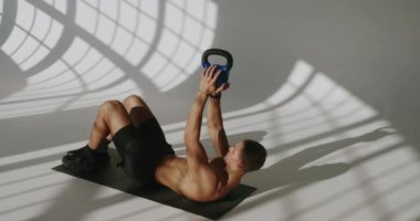 Sporcu adam spor paspası üzerinde egzersiz yapıyor ve stüdyoda karın kasları çalışması için kettlebell 'i elinde tutuyor. Kaslı vücutlu genç erkek ağırlıklarla antrenman yapıyor..