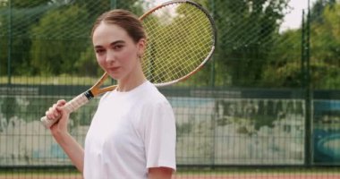 Tenis kortunun önünde duran beyaz tişörtlü güzel bir kadın elinde raketle kameraya bakıyor ve gülümsüyor. Güzel bayan sporcunun portresi maça hazır ol..
