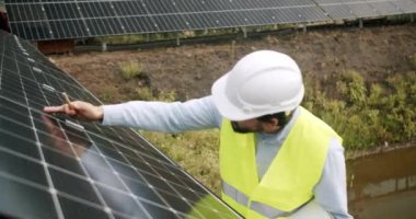 Yelekli ve koruyucu kasklı mühendis dışarıdaki fotovoltaik güç istasyonunda güneş panelini kontrol ediyor. Güneş çiftliğinde çalışan erkek bir uzman. Alternatif enerji kaynakları ve yenilikler kavramı